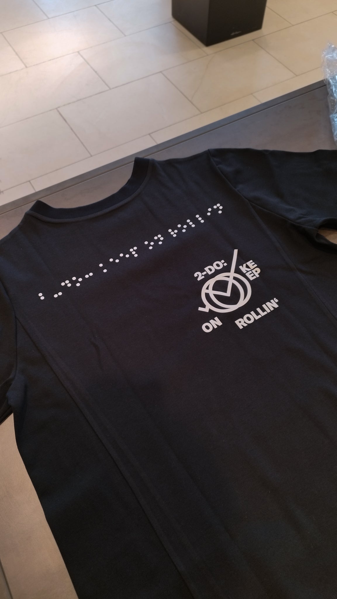 Aufnahme des schwarzen Braille-T-Shirt im Design 2. Die Blindenschrift auf dem oberen Teil des T-Shirts ist in weißer Farbe. Darunter steht rund um ein Icon der Aufdruck 2-Do: Keep on Rollin' in weißer Schrift.