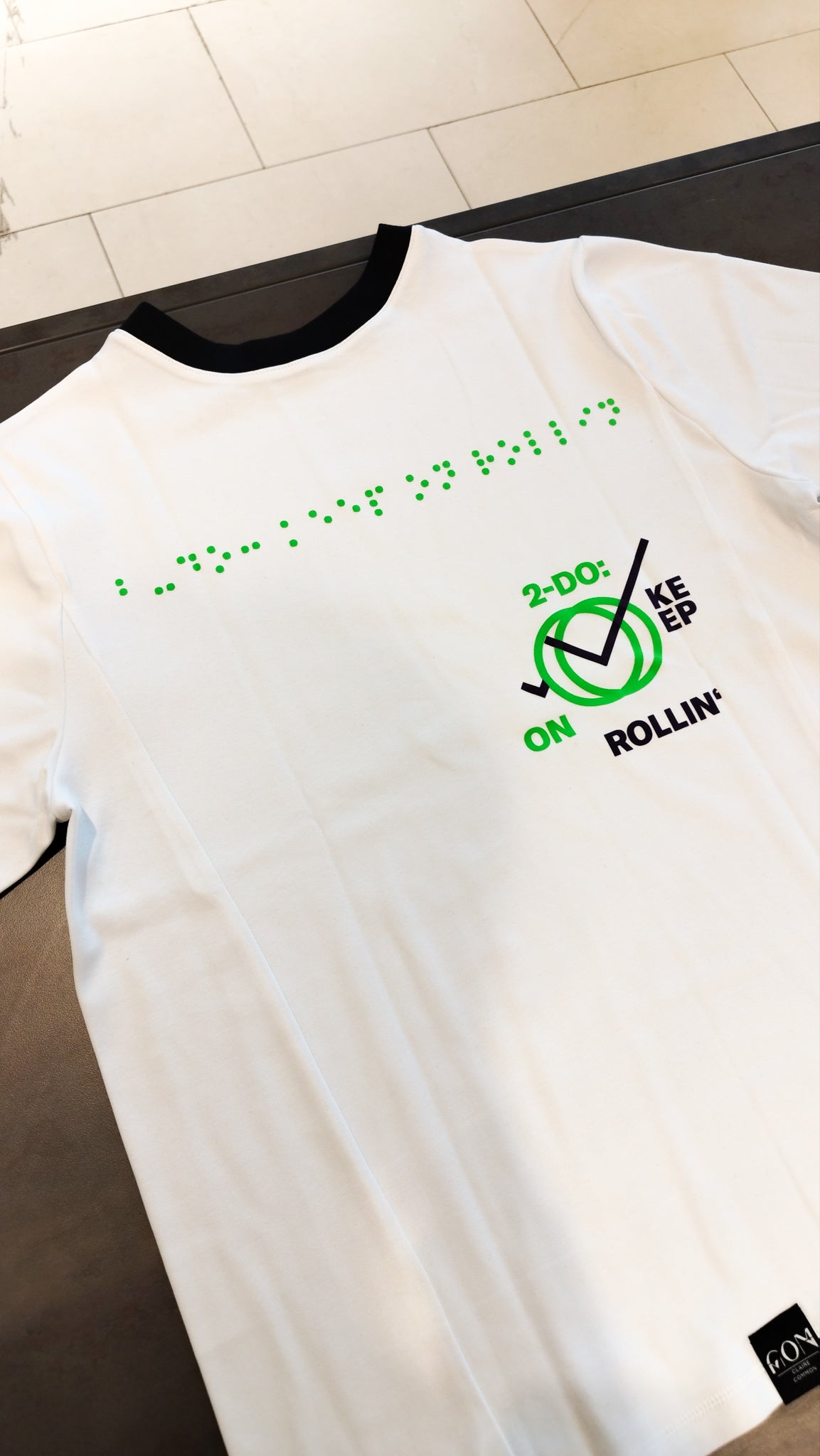 Aufnahme des weißen Braille-T-Shirt im Design 2 mit schwarzer Halsausschnittnaht. Die Blindenschrift auf dem oberen Teil des T-Shirts ist in greller, grüner Farbe. Darunter steht rund um ein Icon der Aufdruck 2-Do: Keep on Rollin' in schwarzer und grell-grüner Schrift.