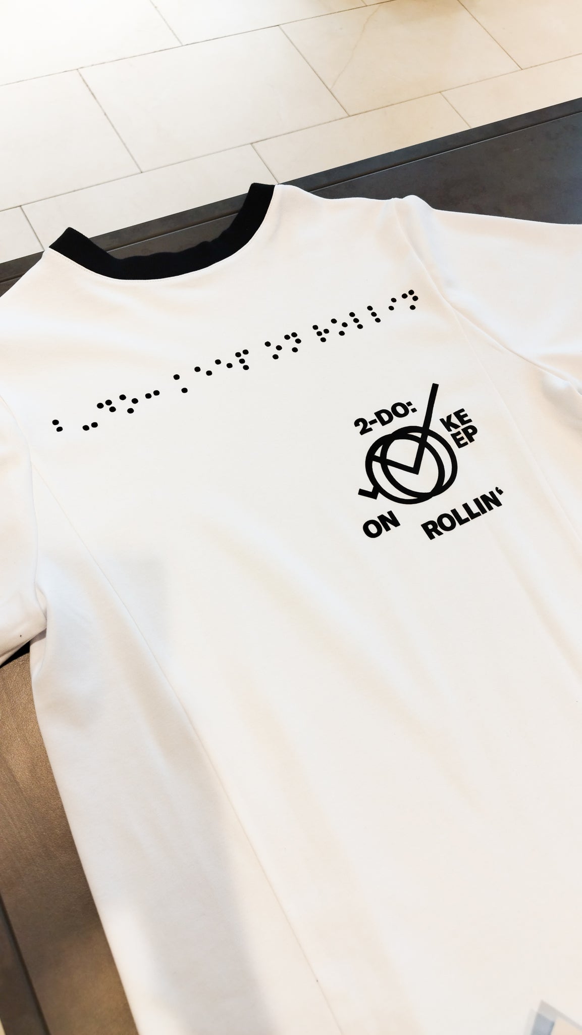 Aufnahme des weißen Braille-T-Shirt im Design 2 mit schwarzer Halsausschnittnaht. Die Blindenschrift auf dem oberen Teil des T-Shirts ist in schwarzer Farbe. Darunter steht rund um ein Icon der Aufdruck 2-Do: Keep on Rollin' in schwarzer und schwarzer Schrift.