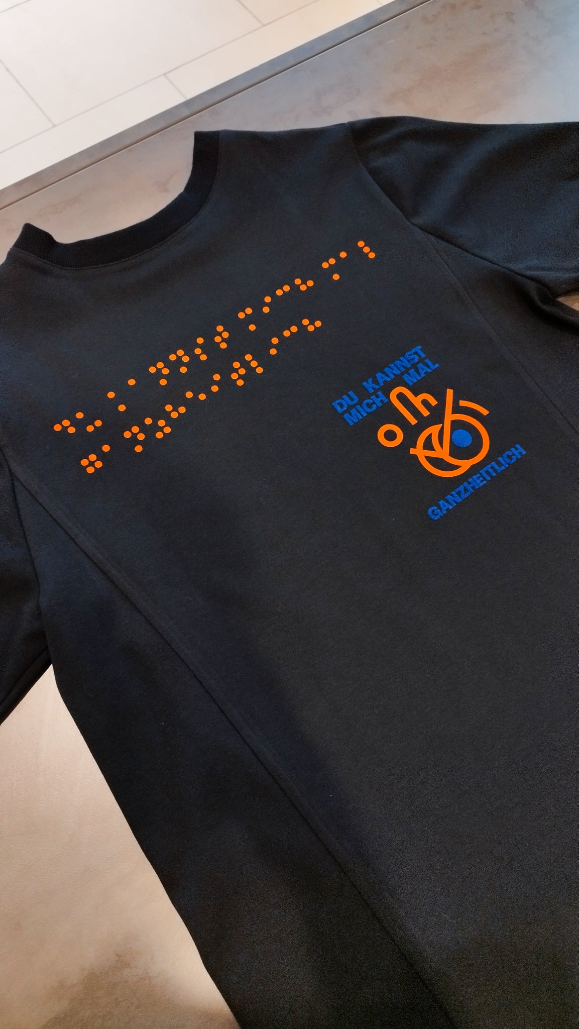 Eine Aufnahme des schwarzen Braille-T-Shirts im Design 3 mit orangener Blindenschrift und dem Aufdruck Du kannst mich mal ganzheitlich! in marineblauer Schrift mit einem orangefarbenen Icon.