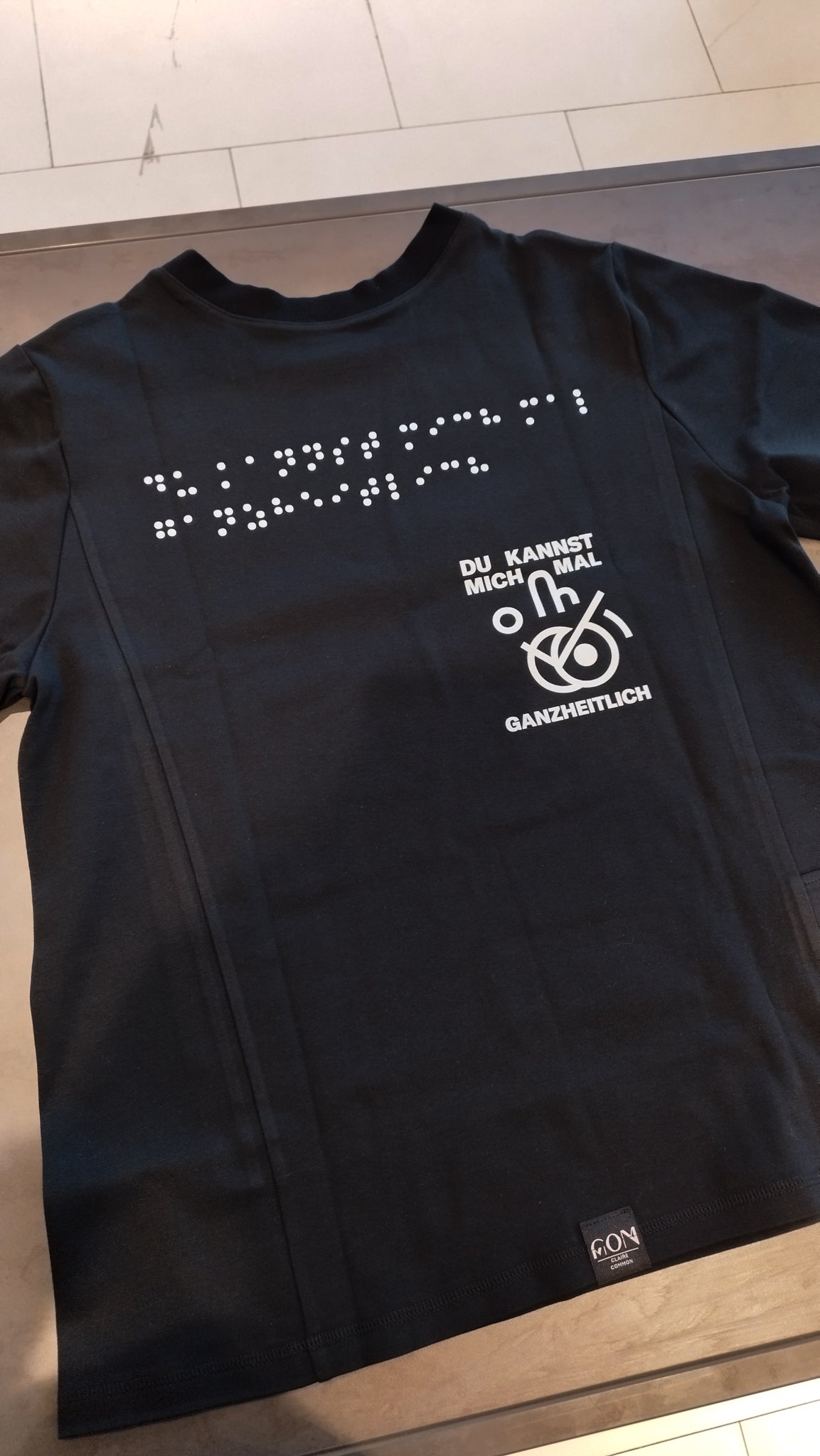 Aufnahme des schwarzen Braille-T-Shirts im Design 3 mit weißer Blindenschrift und dem weißen Aufdruck Du kannst mich mal ganzheitlich! mit einem weißen Icon.