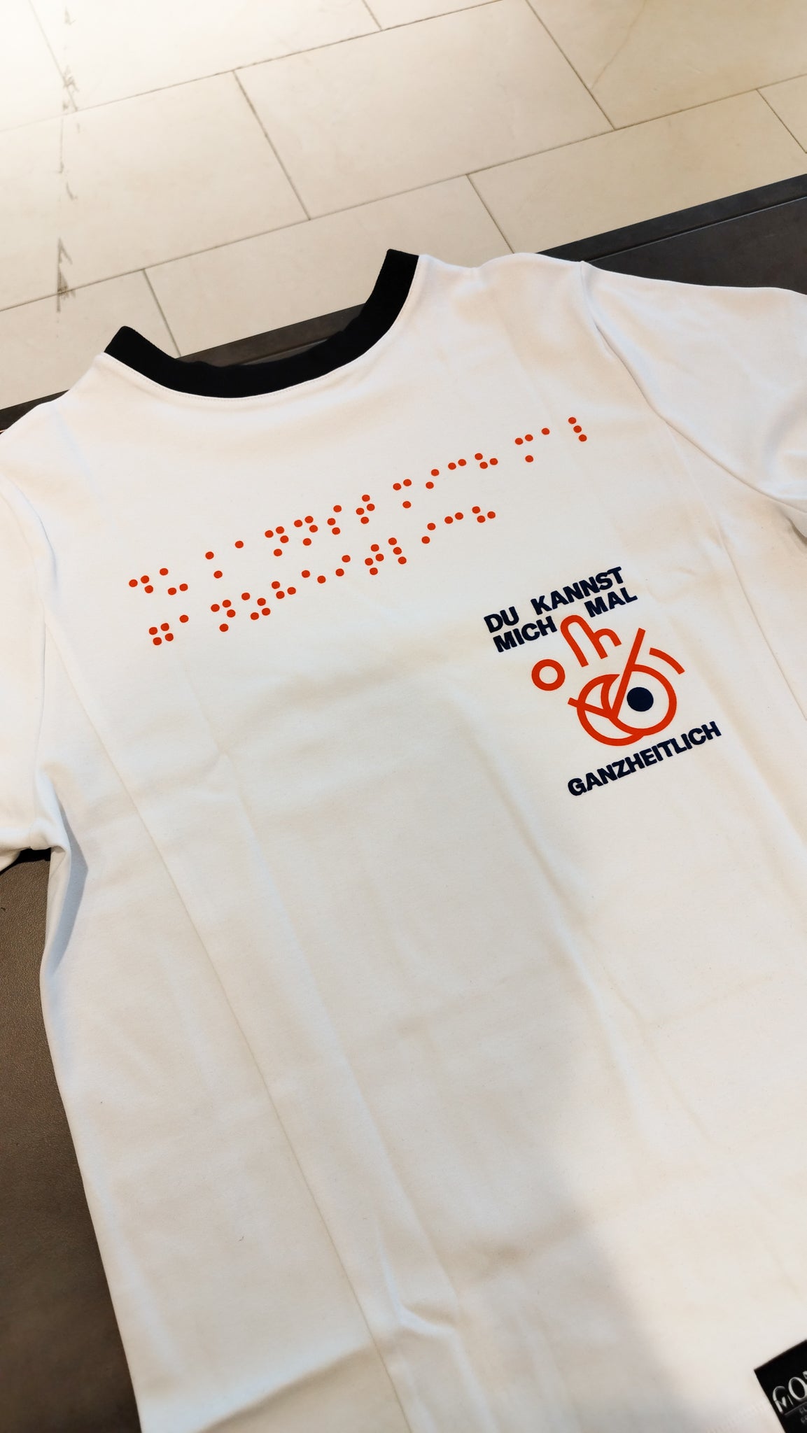 Aufnahme des weißen Braille-T-Shirts im im Design 3 mit einer schwarzen Halsausschnittnaht. Die Blindenschrift auf dem oberen Teil des T-Shirts ist in roter Farbe. Darunter steht in schwarzer Schrift der Schriftzug Du kannst mich mal ganzheitlich! mit einem roten Icon.