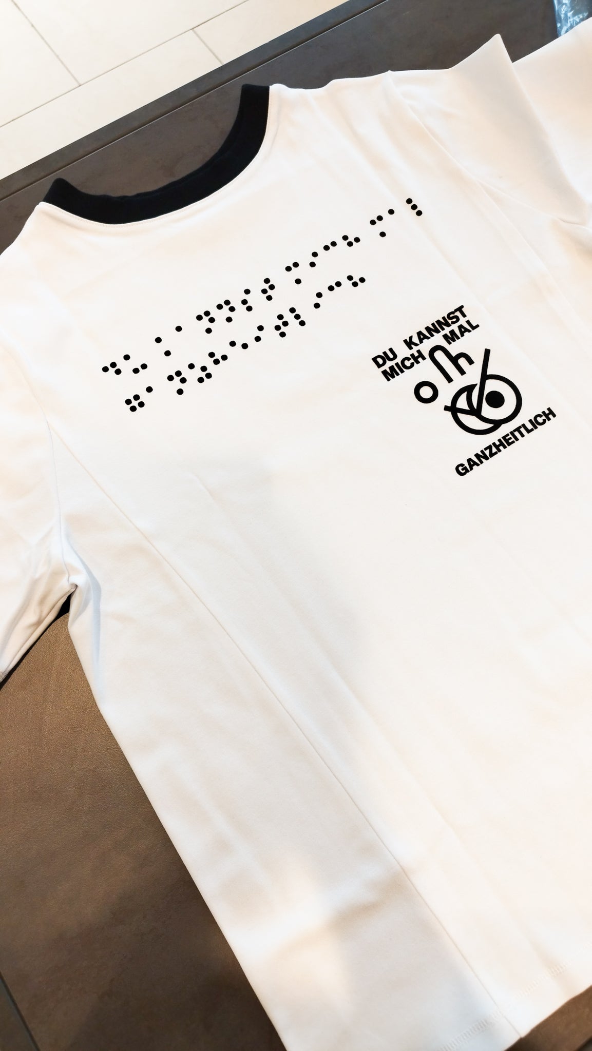 Aufnahme des weißen Braille-T-Shirts im Design 3 mit schwarzer Halsausschnittnaht sowie schwarzer Blindenschrift. Der Spruch und das Icon Du kannst mich mal ganzheitlich sind ebenfalls schwarz.