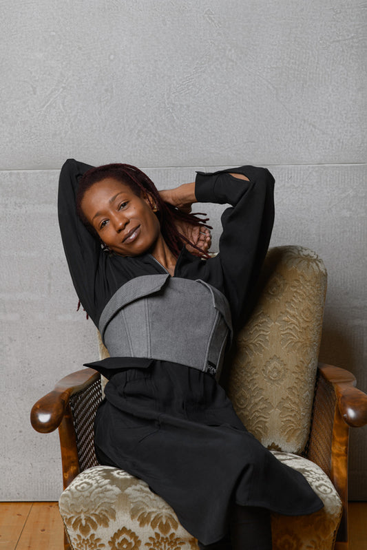 Ein Model sitzt auf einem Stuhl und hat die Arme hinter dem Hinterkopf verschränkt. Es trägt Claire Commons Korsage und lächelt mit seitlich geneigtem Kopf in die Kamera.