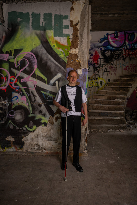 Ein blinder Mann mit einem Blindenstock in seiner rechten Hand steht vor einer Grafitti-Wand. Er schaut frontal in Richtung Kamera und lächelt dabei leicht.