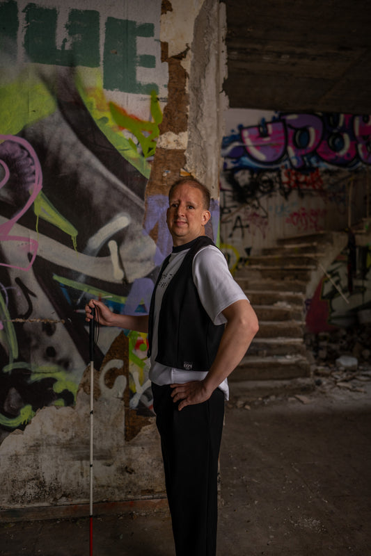Ein blinder Mann mit einem Blindenstock in seiner rechten Hand steht vor einer Grafitti-Wand. Seinen linken Arm hat er lässig in die Hüfte gestemmt. Er schaut in Richtung Kamera und lächelt leicht.