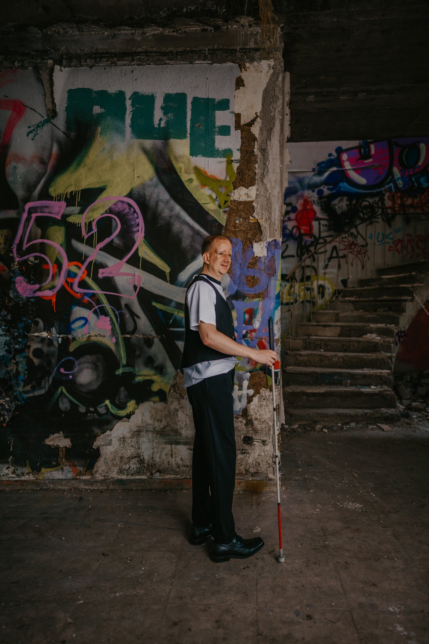 Ein blinder Mann mit einem Blindenstock in seiner rechten Hand steht vor einer Grafitti-Wand. Er schaut an den rechten Bildrand und lächelt leicht.