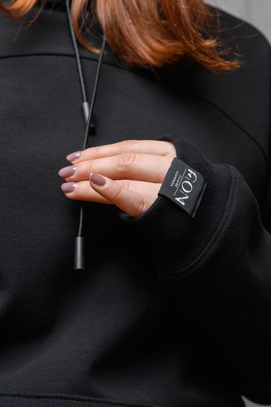 Eine Nahaufnahme des Etiketts von Claire Common am Ärmelsaum des Unisex-Hoodies. Eine Frau hält die Kordeln des Pullis dabei vor ihrem Oberkörper in die Kamera.