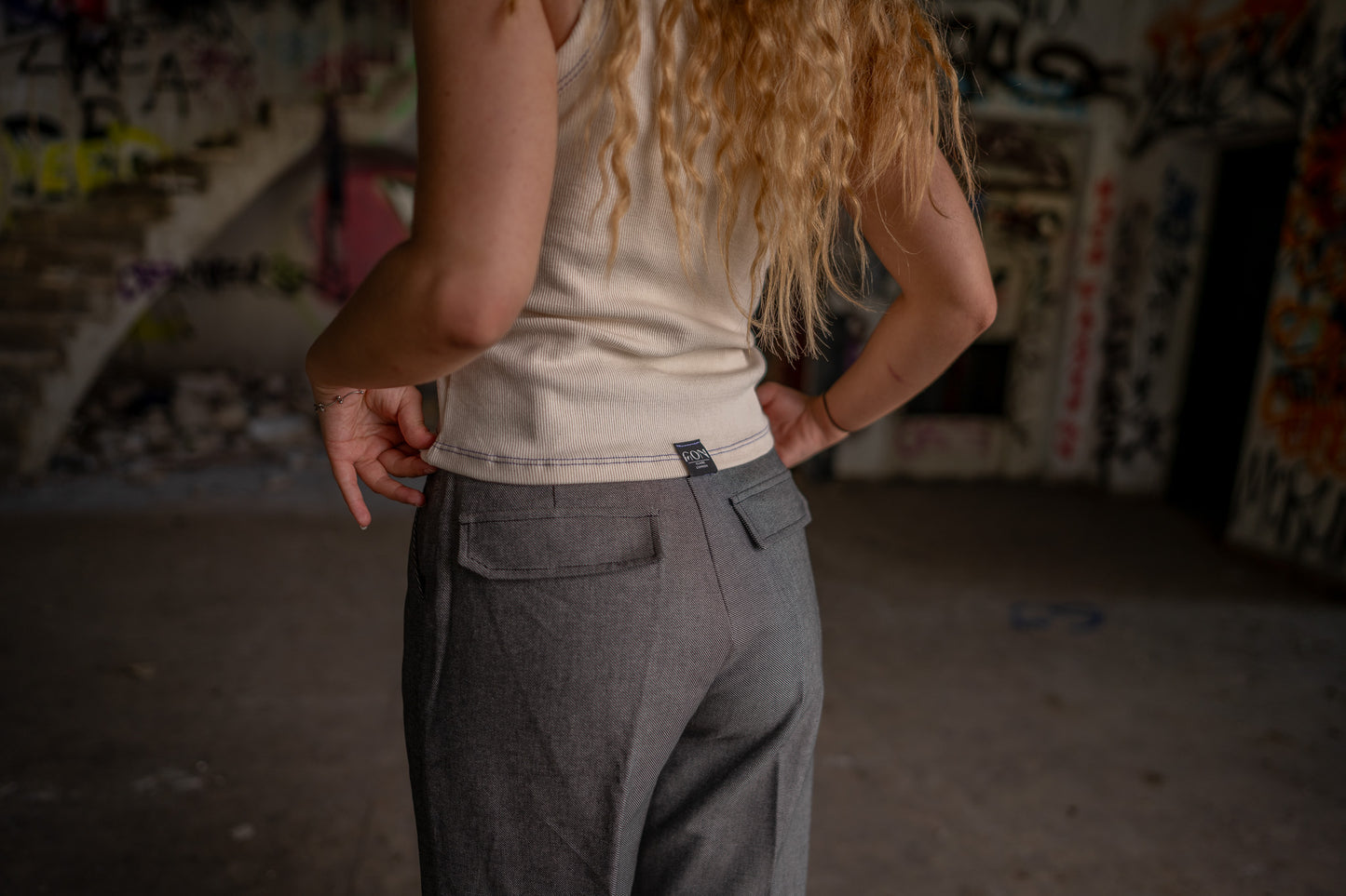 Rückenaufnahme vom hellen Tanktop mit schwarzer Naht an einem Model. Das Model trägt eine graue Hose und darüber das helle Tanktop. In der Mitte des unteren Saums ist das Etikett von Claire Common genäht.