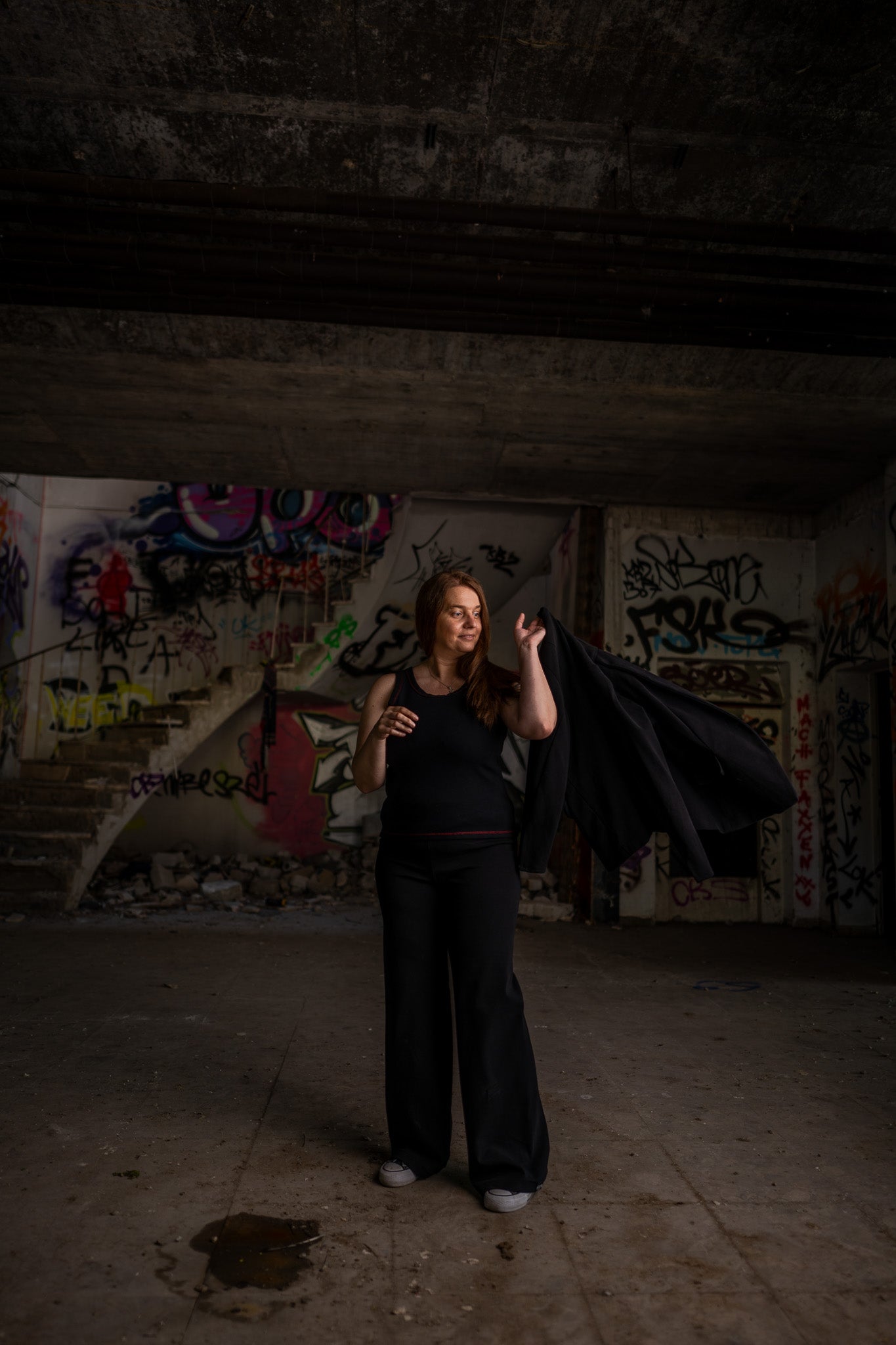 Junge Frau vor einer Graffiti-Wand, sie trägt Claires schwarzes Tanktop und schwingt selbstbewusst eine Jacke über ihre linke Schulter.