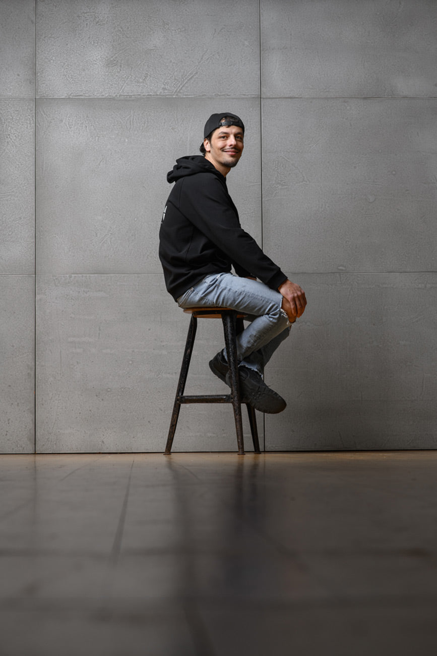 Ein Mann sitzt seitlich vor einer grauen Wand auf einem Stuhl und schaut lächelnd in die Kamera. Er trägt den Unisex-Hoodie von Claire Common sowie eine Cap und lächelt in die Kamera.