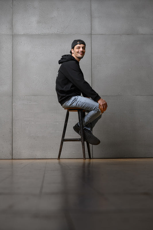 Ein Mann sitzt seitlich vor einer grauen Wand auf einem Stuhl und schaut lächelnd in die Kamera. Er trägt den Unisex-Hoodie von Claire Common sowie eine Cap und lächelt in die Kamera.