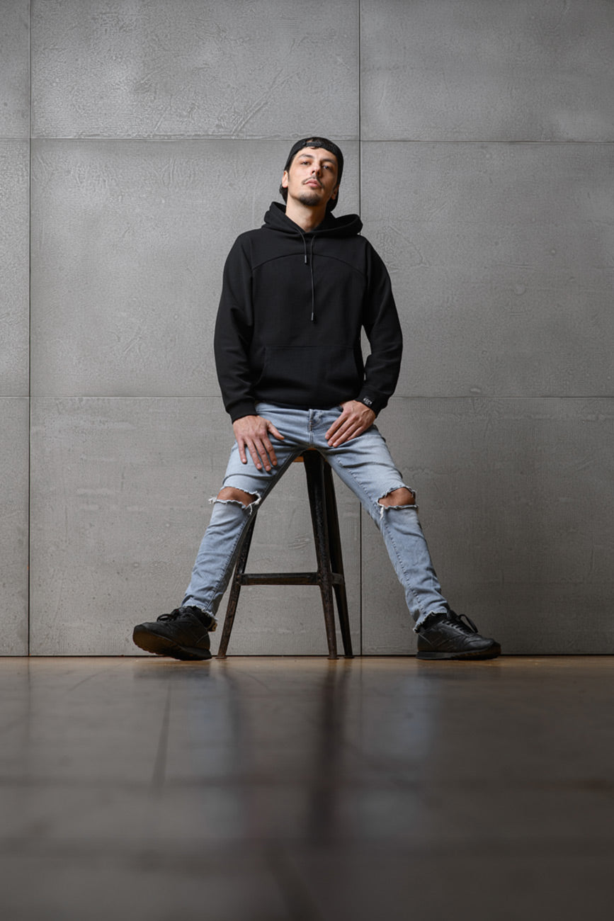 Ein Mann sitzt auf einem Stuhl vor einer grauen Wand und blickt gerade in die Kamera. Er trägt den Unisex-Hoodie von Claire Common, den man von vorne sehen kann.