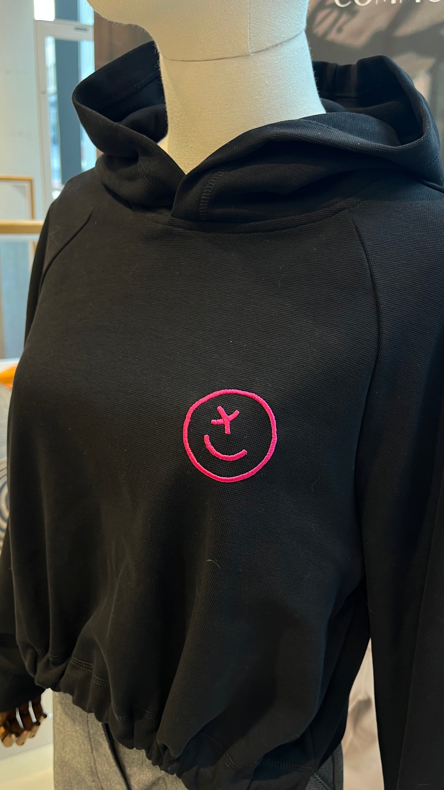 Auf einer Schneider-Puppe ist eine Nahaufnahme des gerafften Hoodies mit pinkem gestickten Smiley zu sehen.