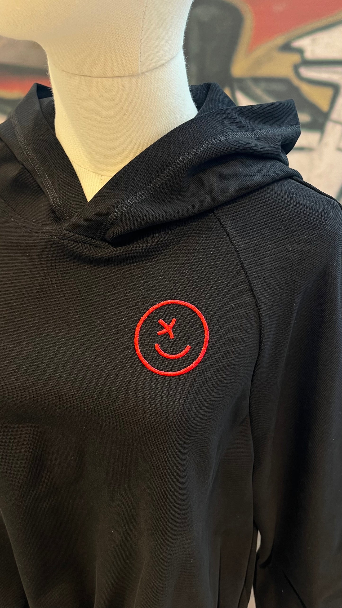 Auf einer Schneider-Puppe ist eine Nahaufnahme des gerafften Hoodies mit rotem gestickten Smiley zu sehen.
